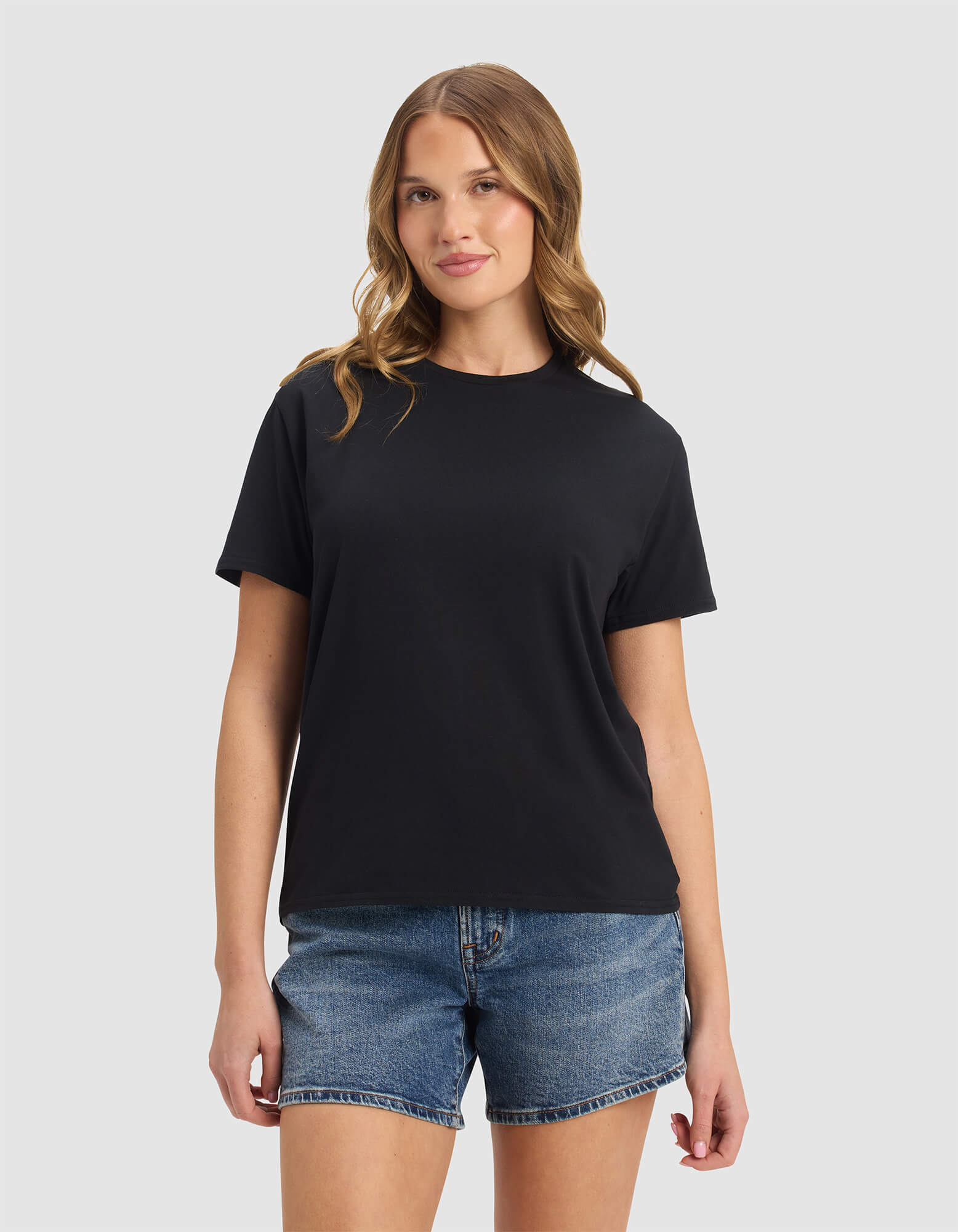 A woman with wavy brown hair wears the Cozy Earth Women's Go-To Tee in Jet Black and blue denim shorts, standing against a light gray background and smiling slightly.