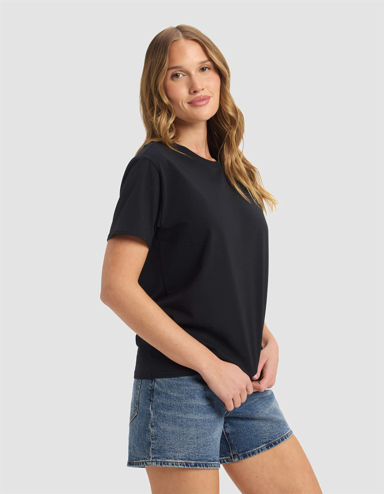 A woman with long, wavy hair models the Cozy Earth Women's Go-To Tee in black with denim shorts, standing against a light gray background and smiling slightly at the camera.