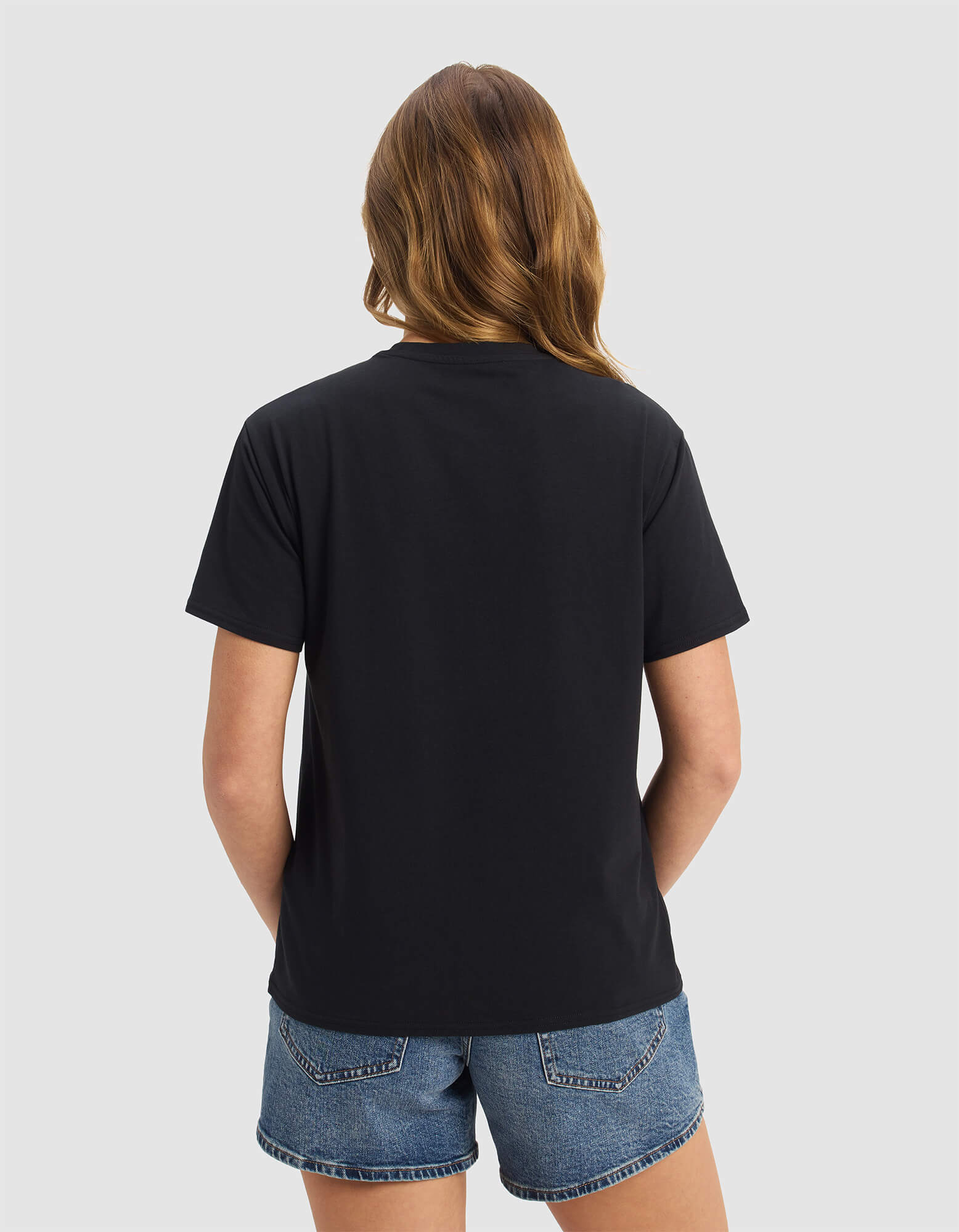 A woman with long, light brown hair stands with her back to the camera, wearing the Cozy Earth Women's Go-To Tee in black with denim shorts against a light gray background.
