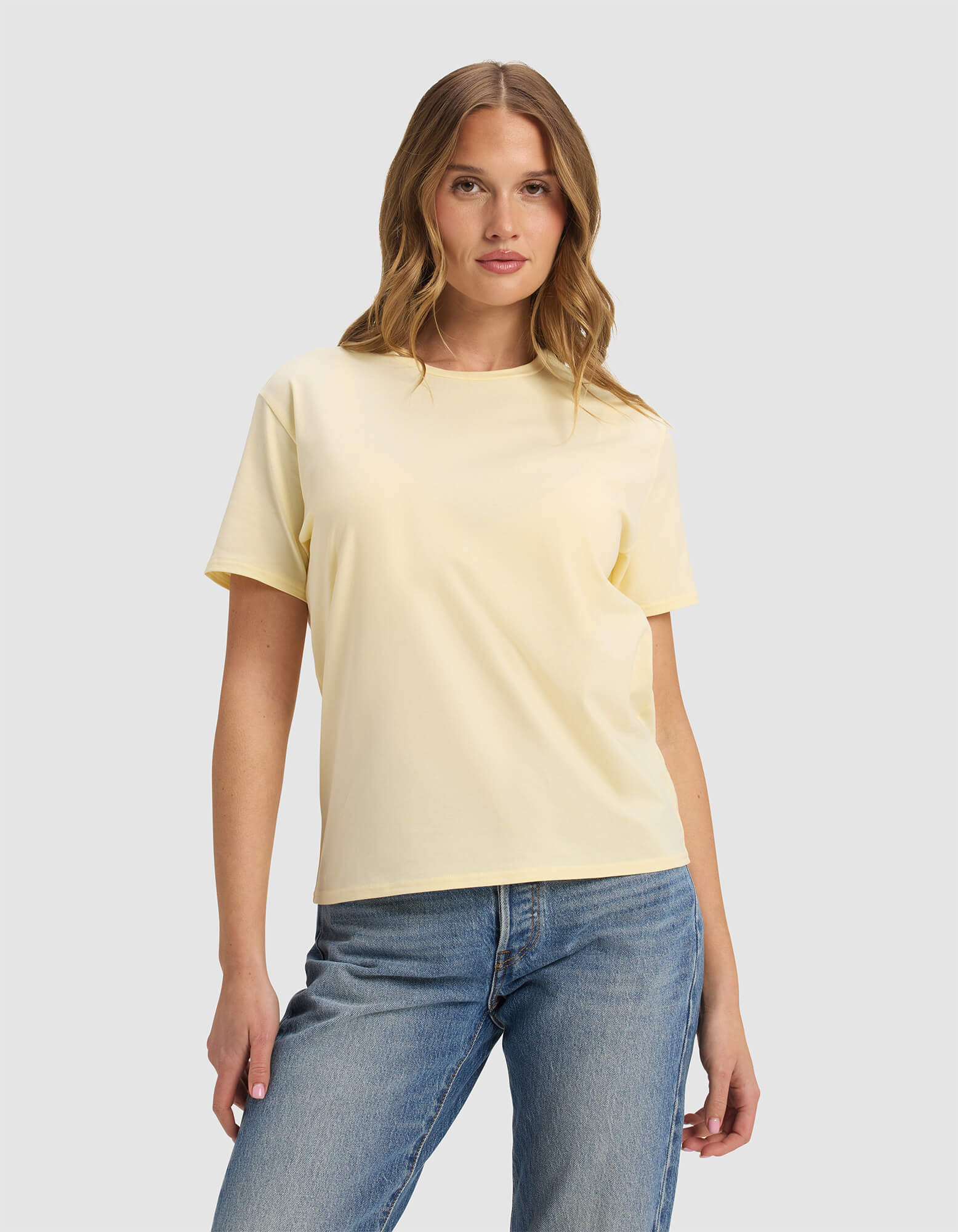 A woman with wavy light brown hair wears the Cozy Earth Women's Go-To Tee in Meringue and blue jeans, standing against a plain light gray background.