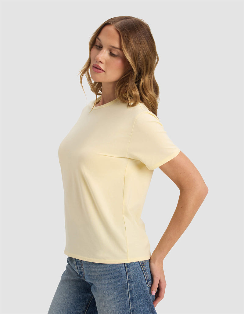 A woman with wavy, light brown hair wears the Cozy Earth Women's Go-To Tee in pale yellow and blue jeans. She stands sideways, eyes closed, arms slightly behind her against a plain light background.