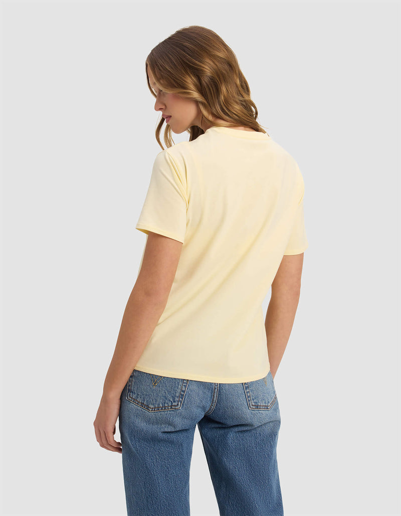 A woman with wavy light brown hair models the Cozy Earth Women's Go-To Tee in pale yellow, paired with blue jeans, as she stands facing away and glances over her left shoulder against a plain light gray background.
