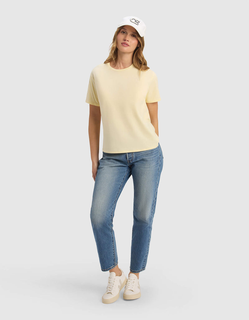 A woman stands with one hand in her pocket, wearing the Cozy Earth Women's Go-To Tee in pale yellow, a white cap, blue jeans, and white sneakers against a plain light gray background.