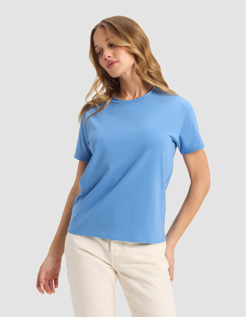 A woman with wavy light brown hair models the Cozy Earth Women's Go-To Tee in Sailor, paired with off-white pants. She stands against a plain light grey background, looking slightly down with a relaxed expression.