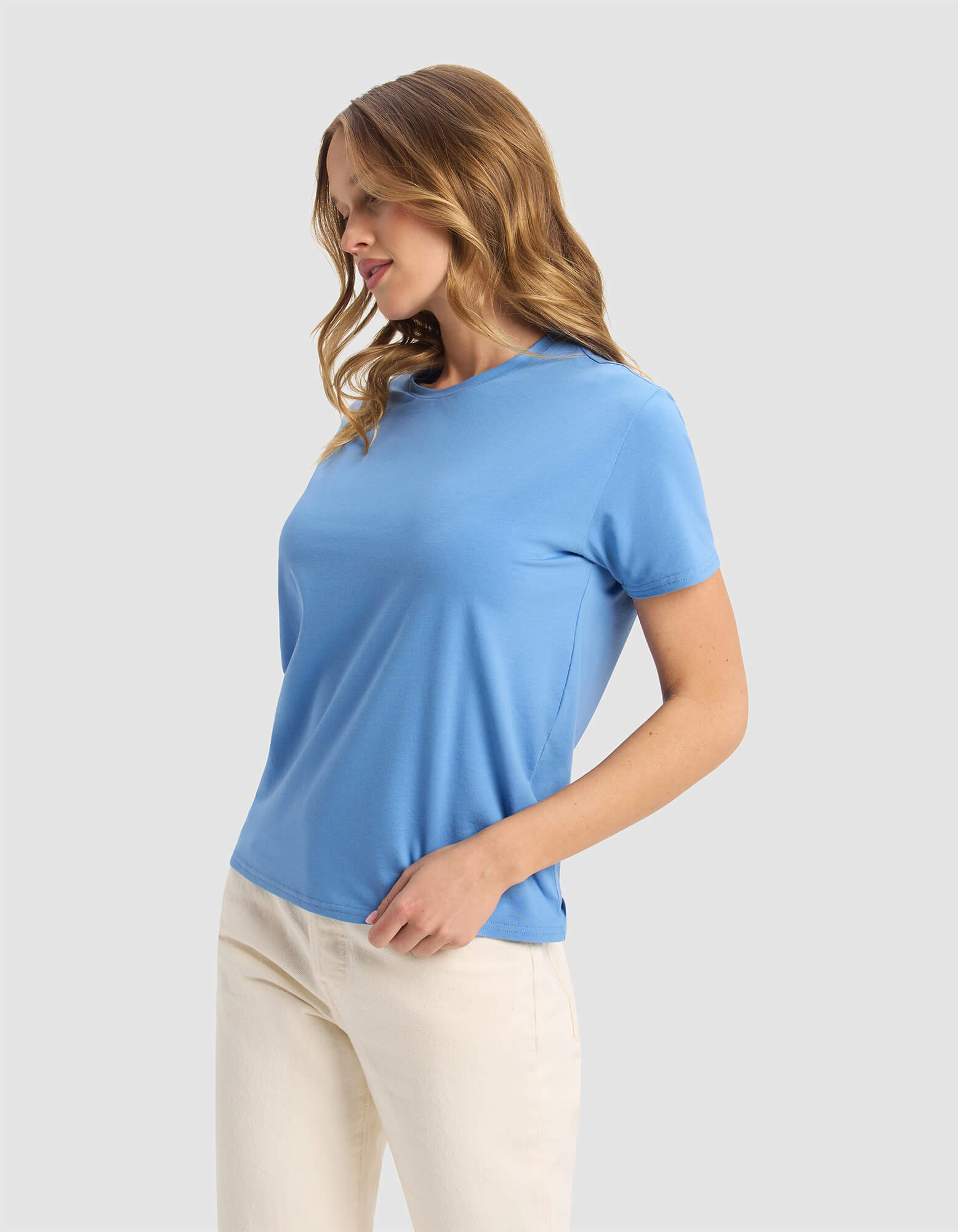 A woman with wavy light brown hair wears the Cozy Earth Women's Go-To Tee in light blue and off-white pants, standing against a plain light grey background with one hand in her pocket, looking down.