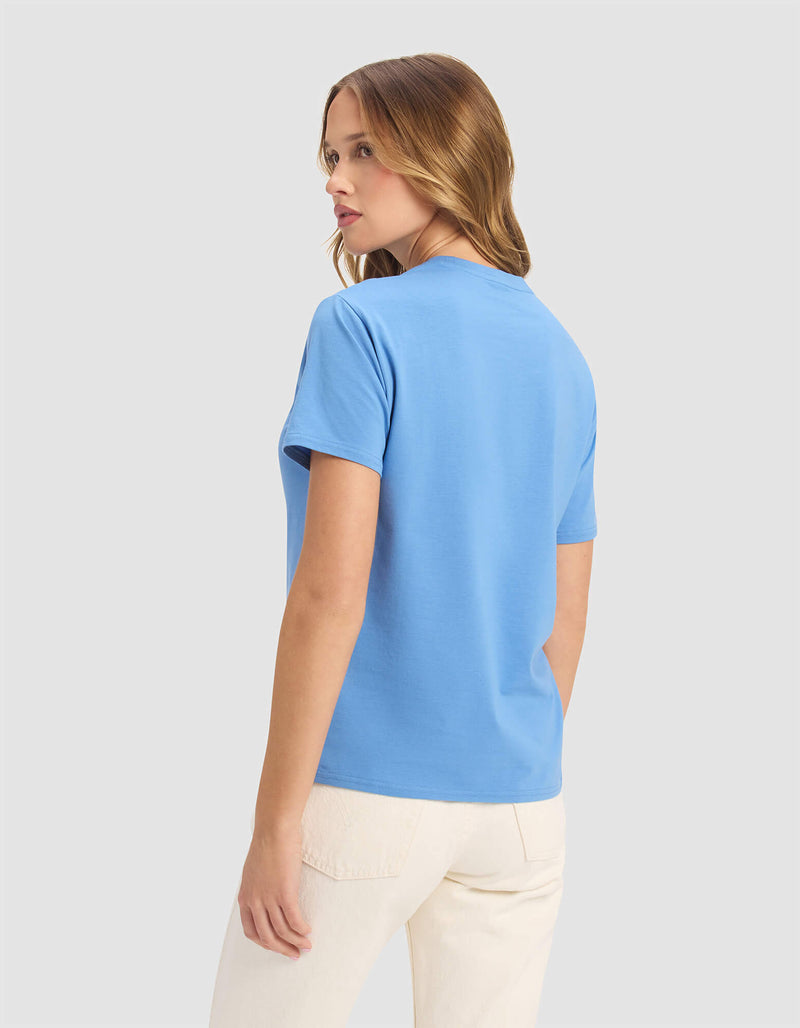 A woman with light brown hair wears the Cozy Earth Women's Go-To Tee in light blue and off-white pants, standing against a plain light gray background, facing away while turning her head slightly to the left.