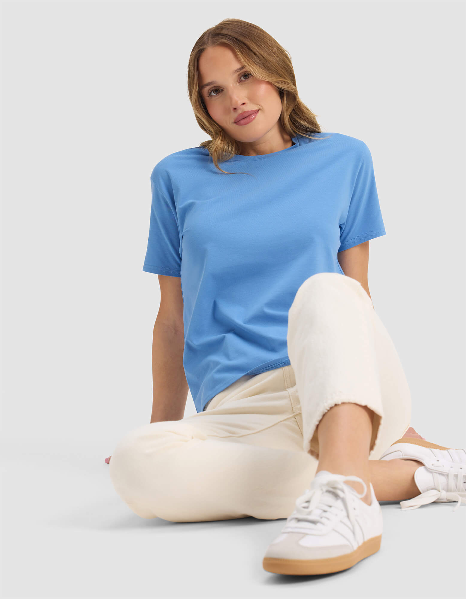 A woman with shoulder-length light brown hair sits against a plain background, looking relaxed with one knee raised. She wears Cozy Earth's Women's Go-To Tee in light blue, paired with cream pants and white sneakers, sporting a slight smile.