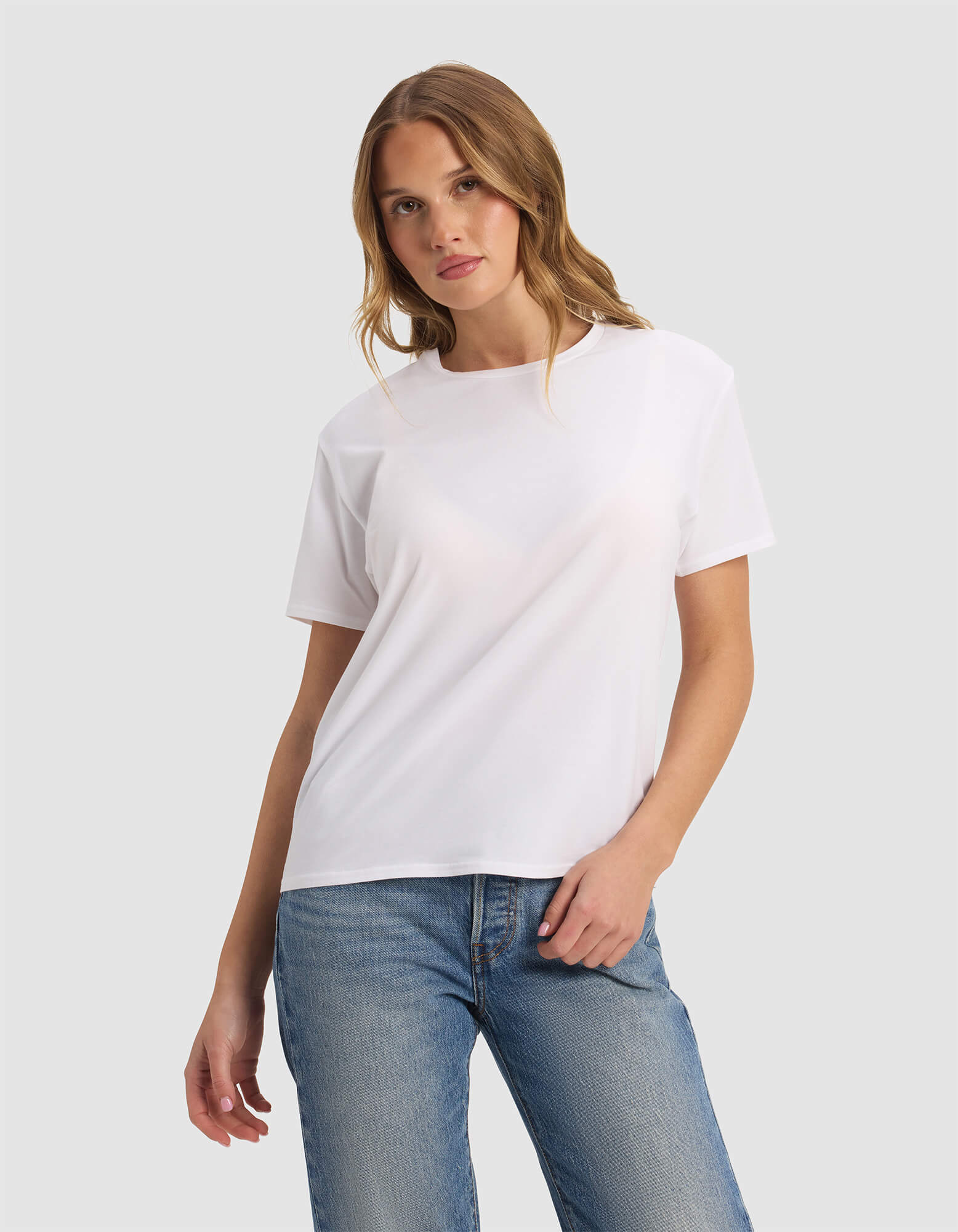 A woman with light brown hair wears the Cozy Earth Women's Go-To Tee in white and blue jeans, standing against a light gray background with one hand in her pocket and looking at the camera.