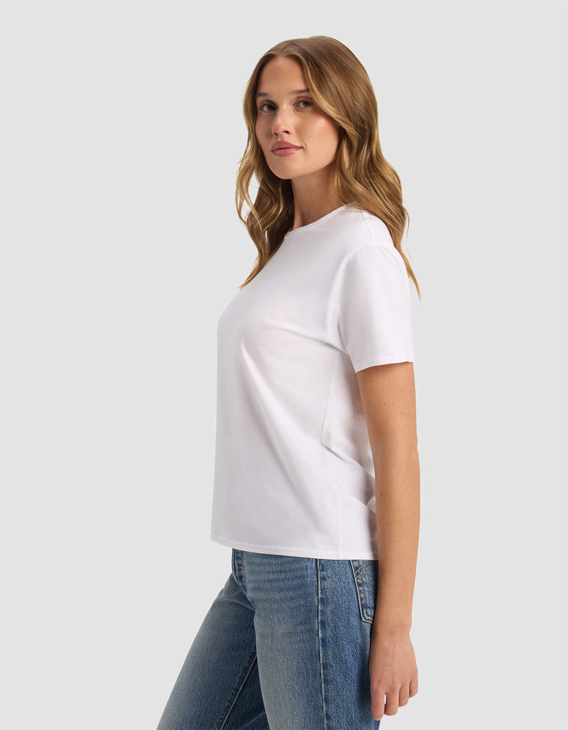 A woman with long, wavy hair wears the Cozy Earth Women's Go-To Tee in white and blue jeans, standing against a light gray background with a neutral expression.