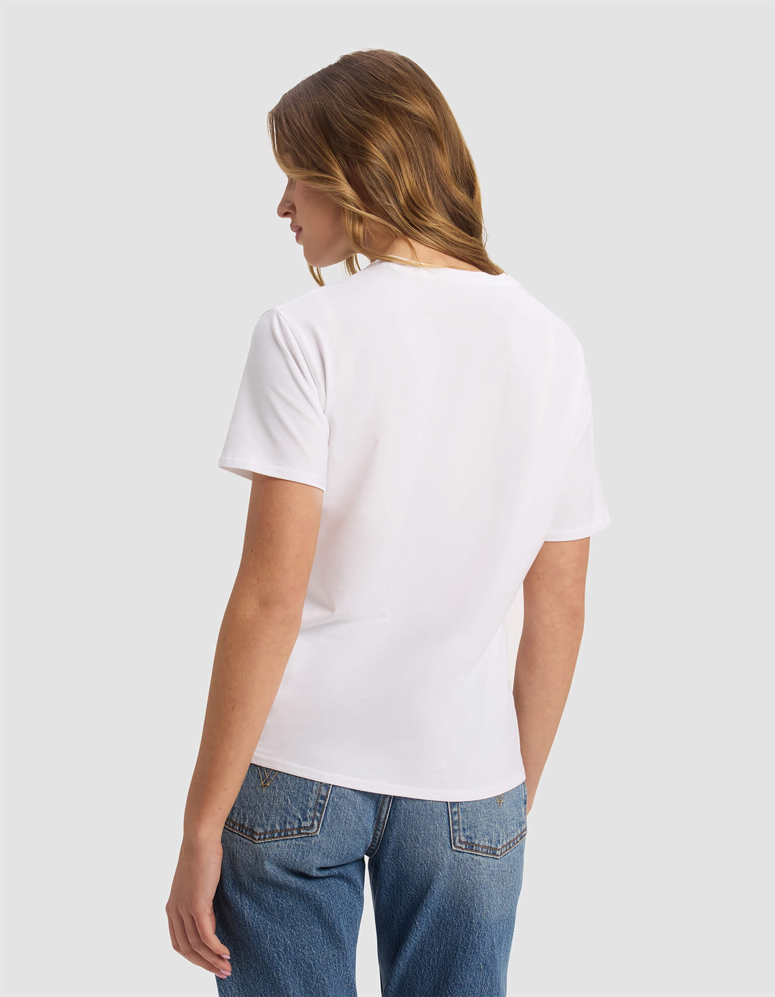 A woman with light brown hair is facing away, wearing the Cozy Earth Women's Go-To Tee in white and blue jeans, set against a light gray background.