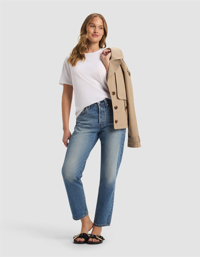 A woman smiles in the Cozy Earth Women's Go-To Tee, paired with blue jeans and black sandals, holding a beige trench coat over her shoulder against a plain light background.