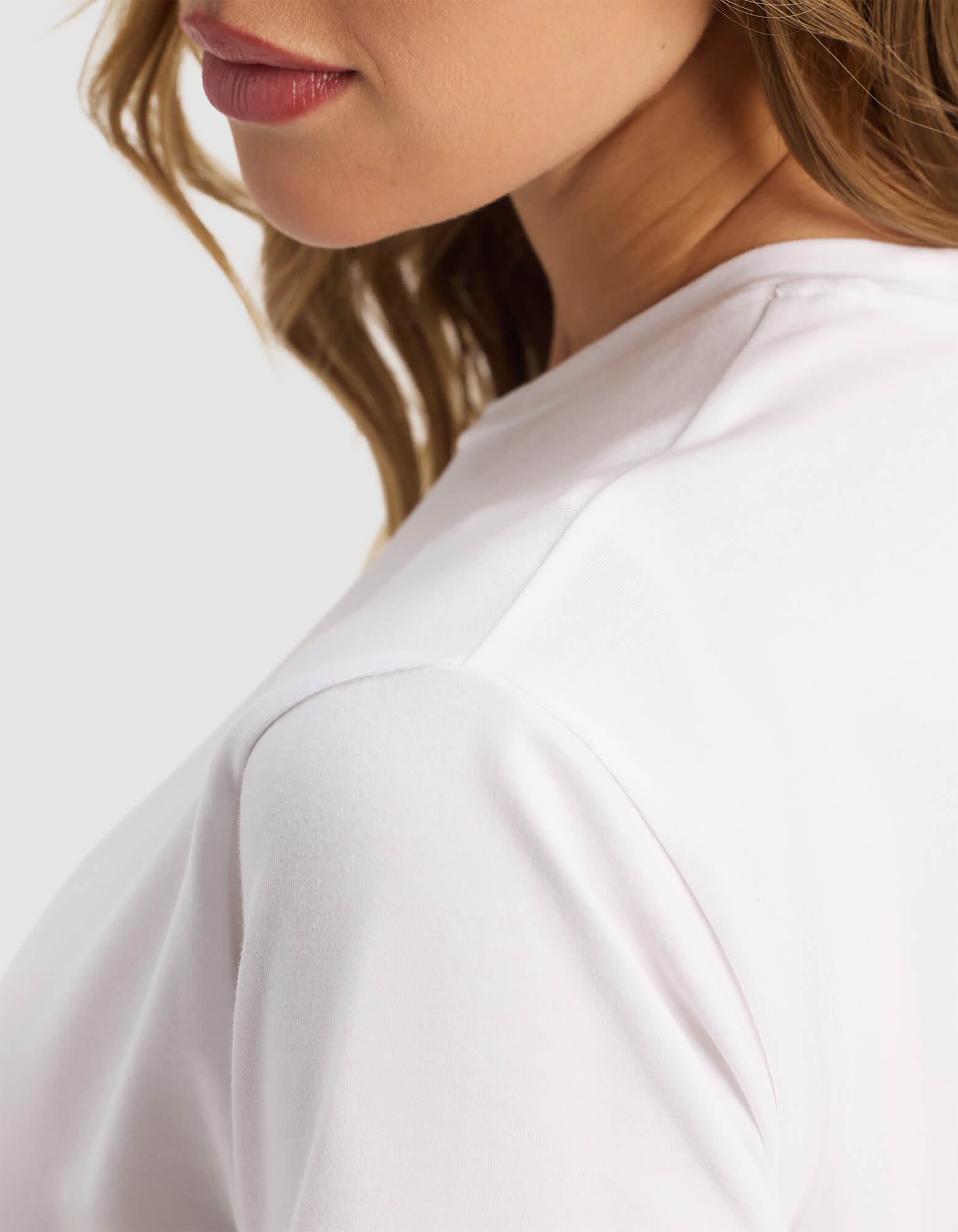 Close-up of a woman wearing the Women's Go-To Tee by Cozy Earth in white, showing her lower face, neck, and shoulder with light brown wavy hair against a plain background.