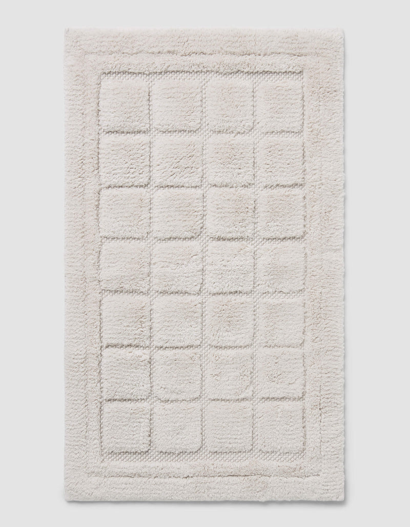 The Cozy Earth Grid Bath Mat is a rectangular, off-white mat with a raised grid design and a thick, fluffy border.