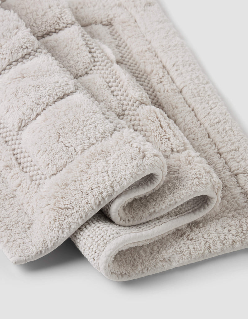 A close-up of two folded Grid Bath Mats by Cozy Earth, featuring a soft, plush surface and geometric texture in beige, displayed on a light gray background.
