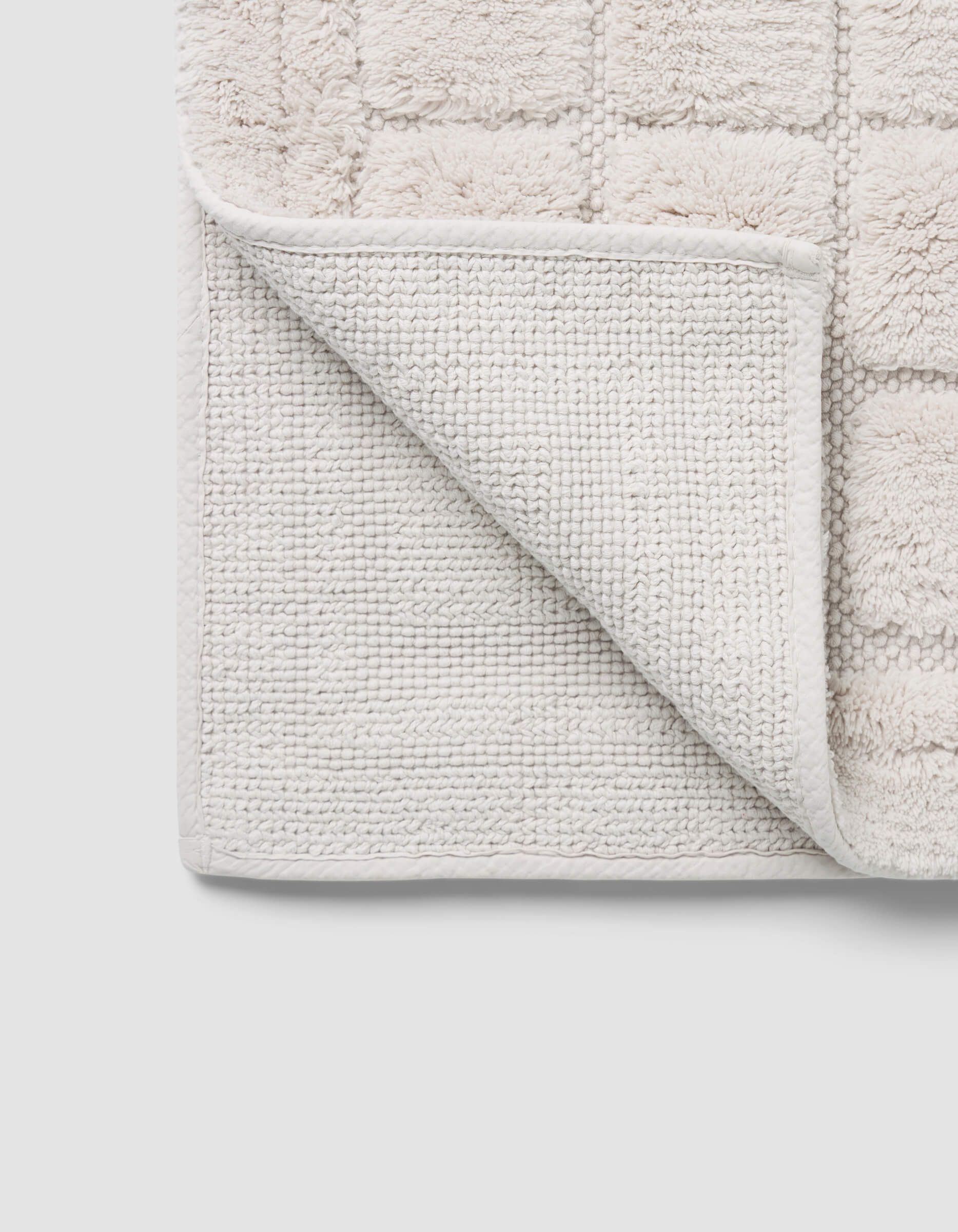 A close-up of the Cozy Earth Grid Bath Mat shows its light color, textured grid pattern, and one corner flipped to reveal the woven underside.|Color:Light Grey