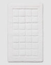 The Cozy Earth Grid Bath Mat is a white, rectangular mat featuring a textured square pattern in the center and a raised border around the edge, shown on a plain background.|Color:White