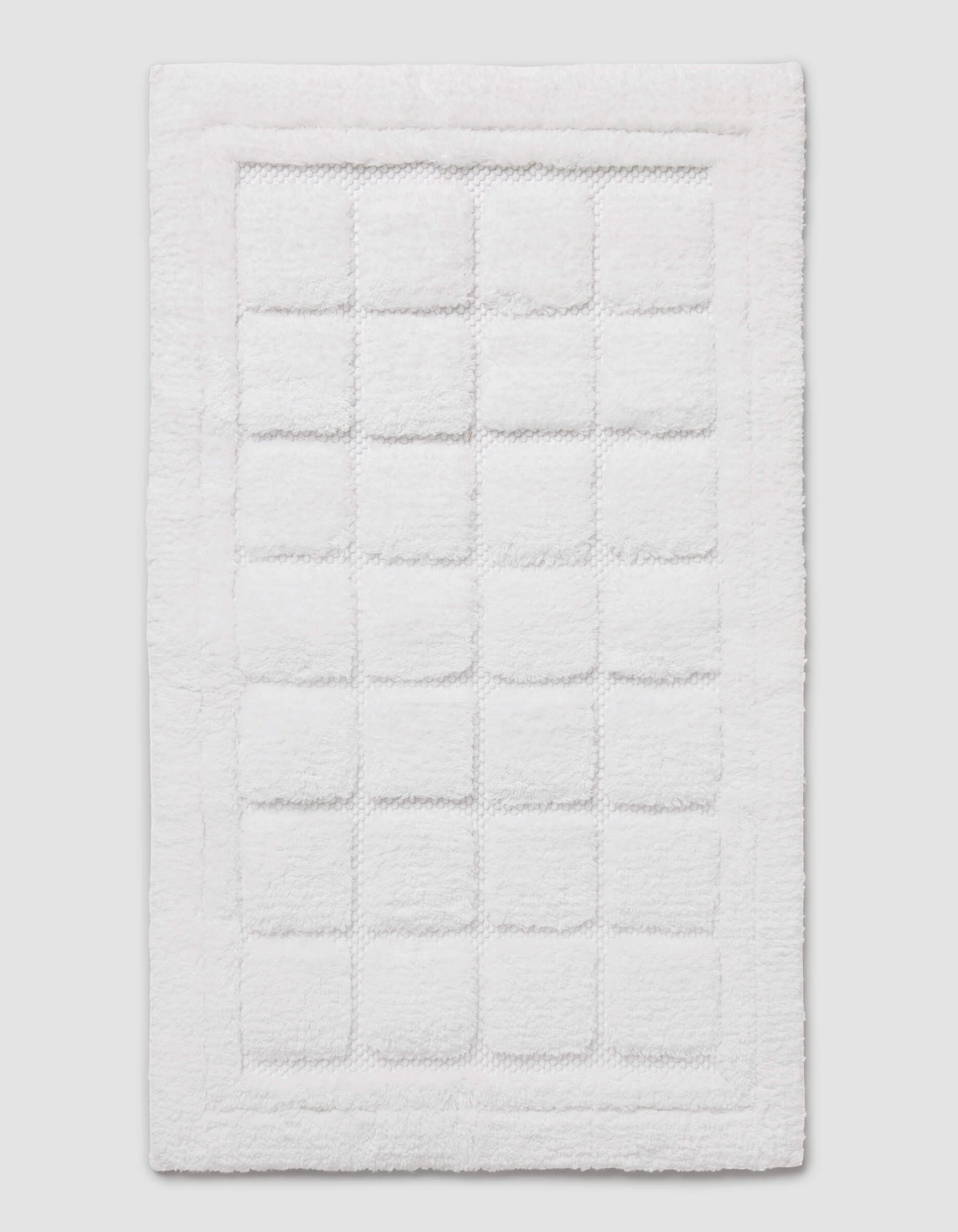 The Cozy Earth Grid Bath Mat is a white, rectangular mat featuring a textured square pattern in the center and a raised border around the edge, shown on a plain background.|Color:White