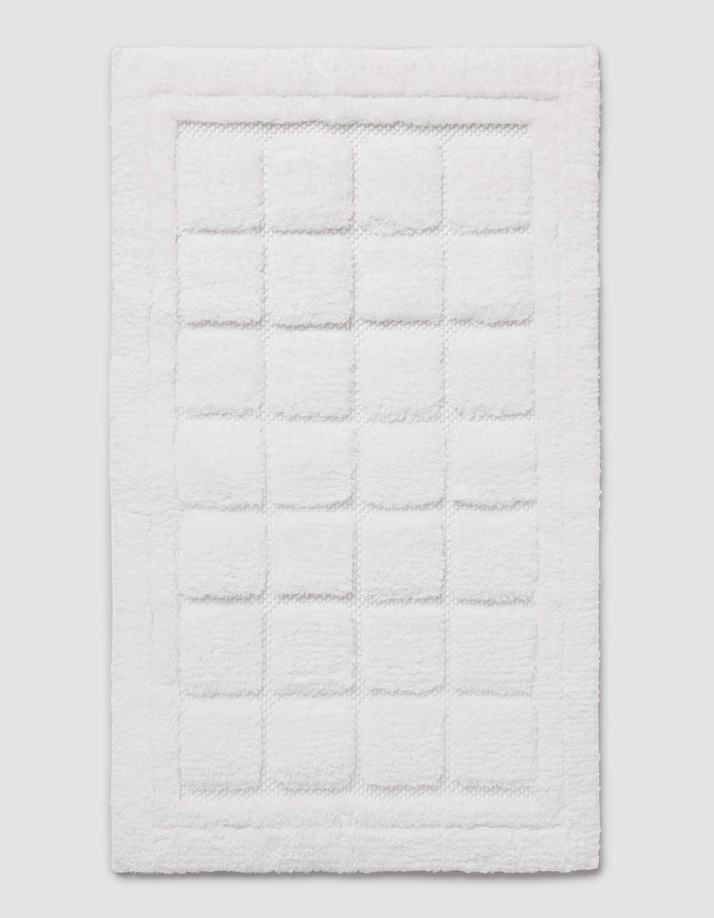 The Cozy Earth Grid Bath Mat is a white, rectangular mat featuring a textured square pattern in the center and a raised border around the edge, shown on a plain background.