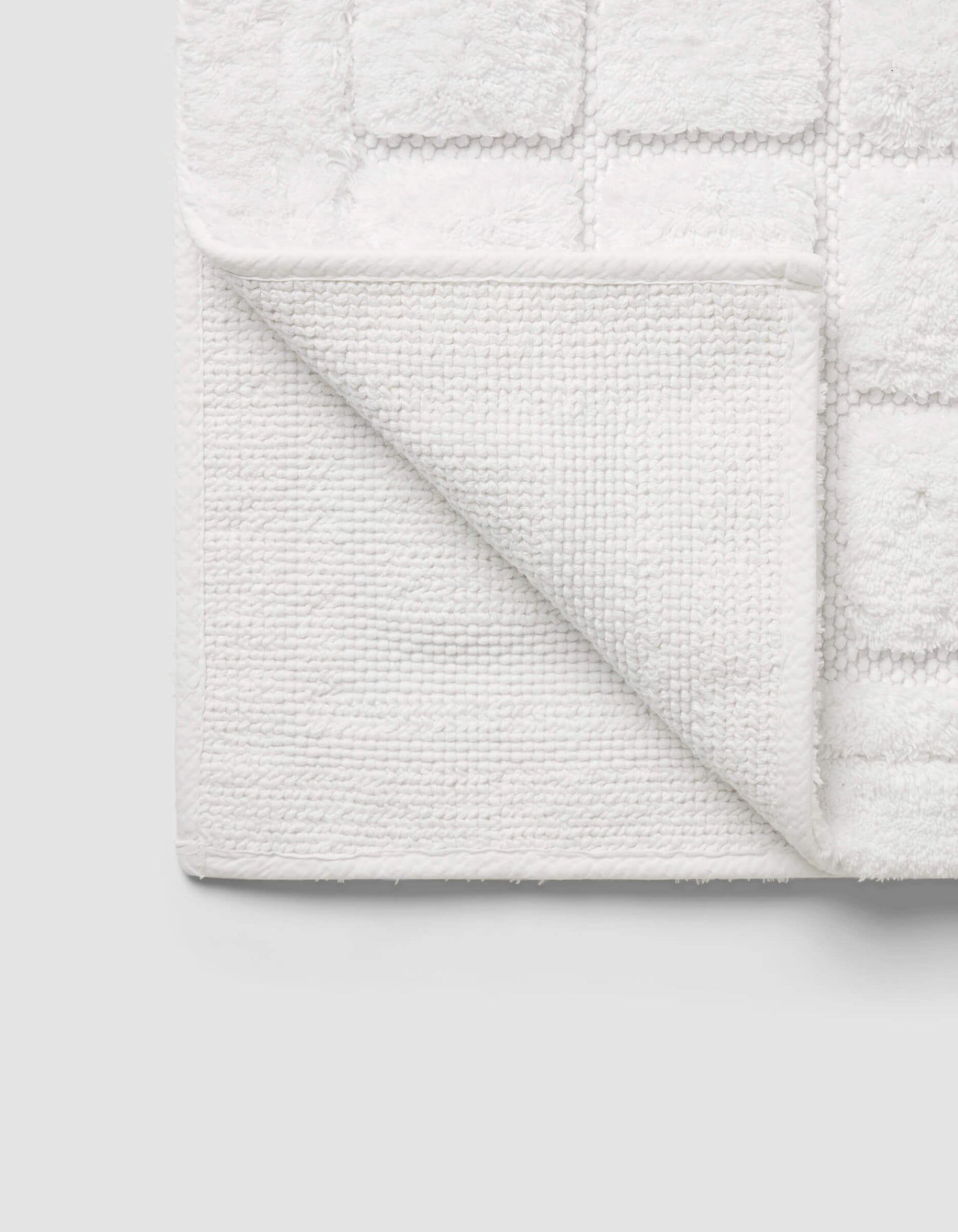 The Cozy Earth Grid Bath Mat, featuring a textured square pattern and woven underside, is displayed with one corner folded on a light gray background.