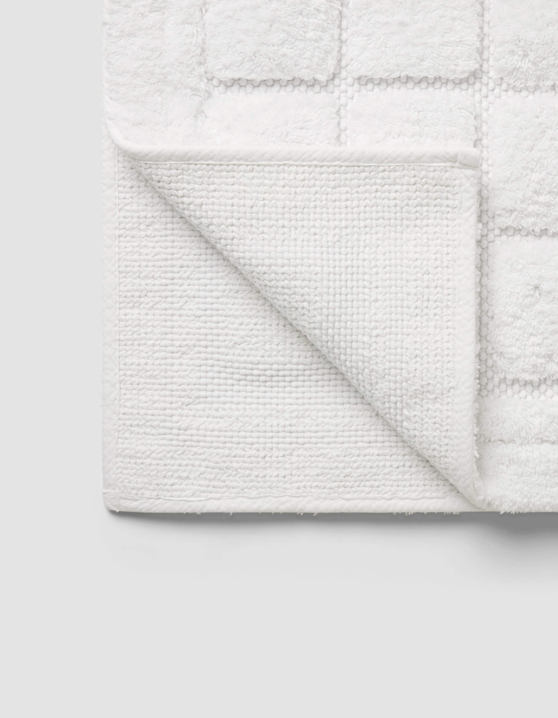 The Cozy Earth Grid Bath Mat, featuring a textured square pattern and woven underside, is displayed with one corner folded on a light gray background.