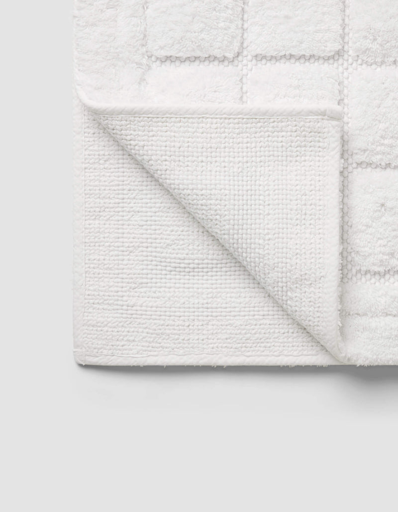 The Cozy Earth Grid Bath Mat, featuring a textured square pattern and woven underside, is displayed with one corner folded on a light gray background.