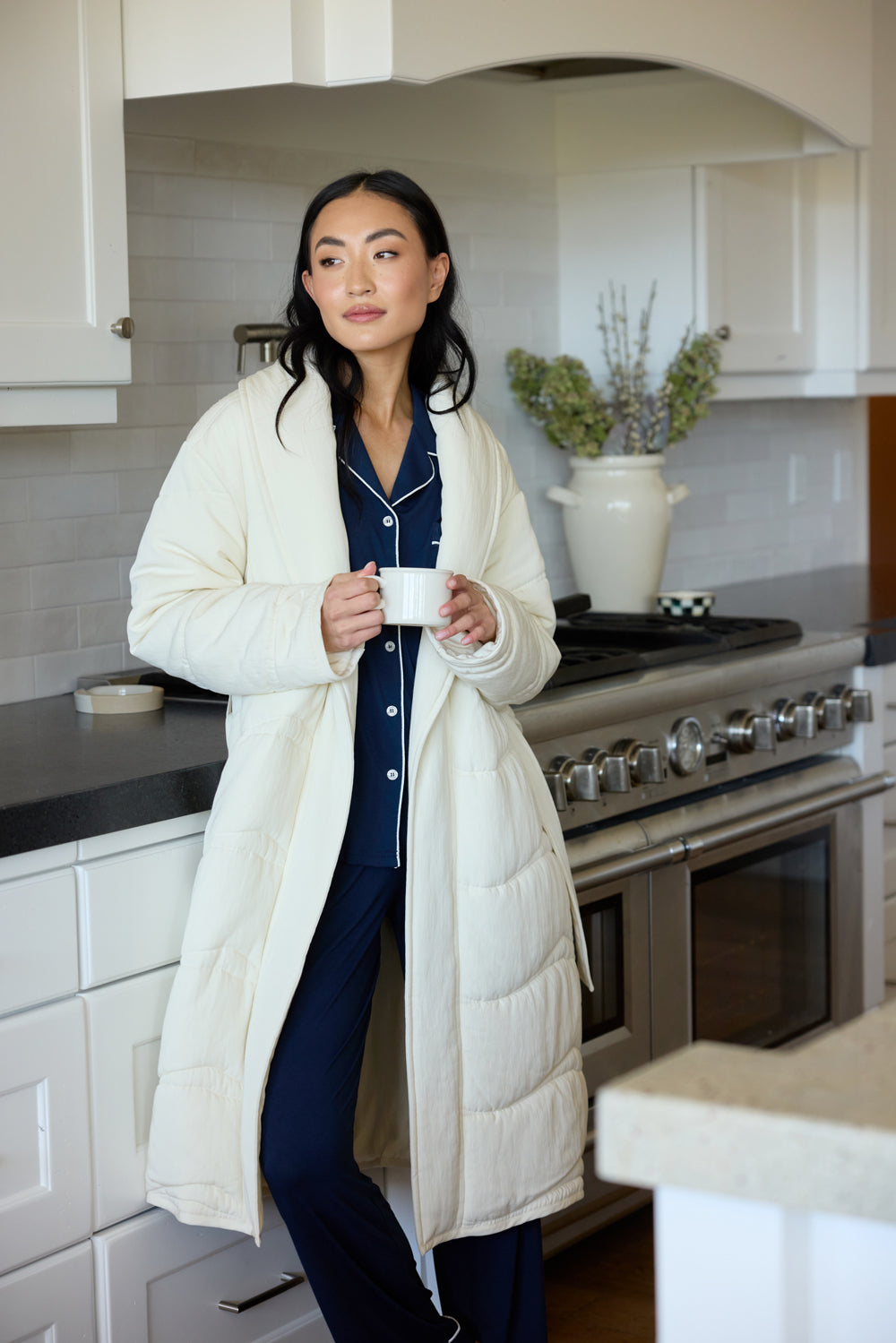 A woman in a kitchen wears Cozy Earth House Coat over her pajamas. She holds a mug.