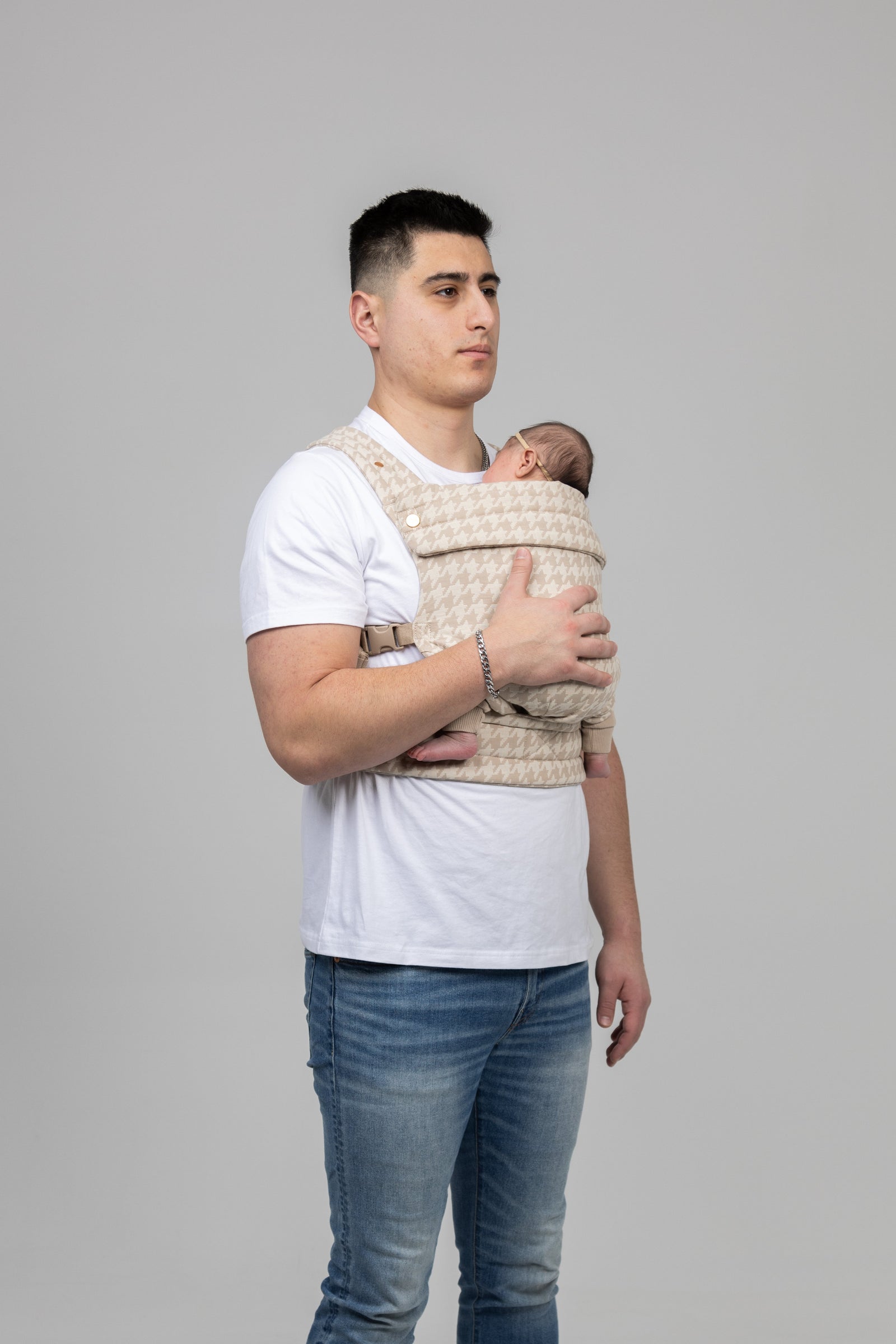 A man in a white t-shirt and jeans stands holding a baby nestled in the Mabē Monarch Carrier - Houndstooth against a plain light gray background. Both look calm and relaxed.