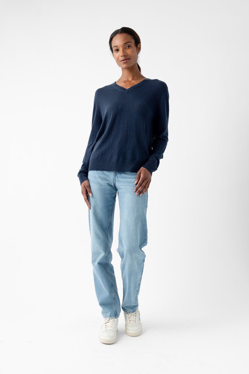 A person stands against a plain white background wearing Cozy Earth's Women's AirKnit V-Neck Sweater in dark blue, light blue jeans, and white sneakers. They are facing forward with their hands at their sides. 