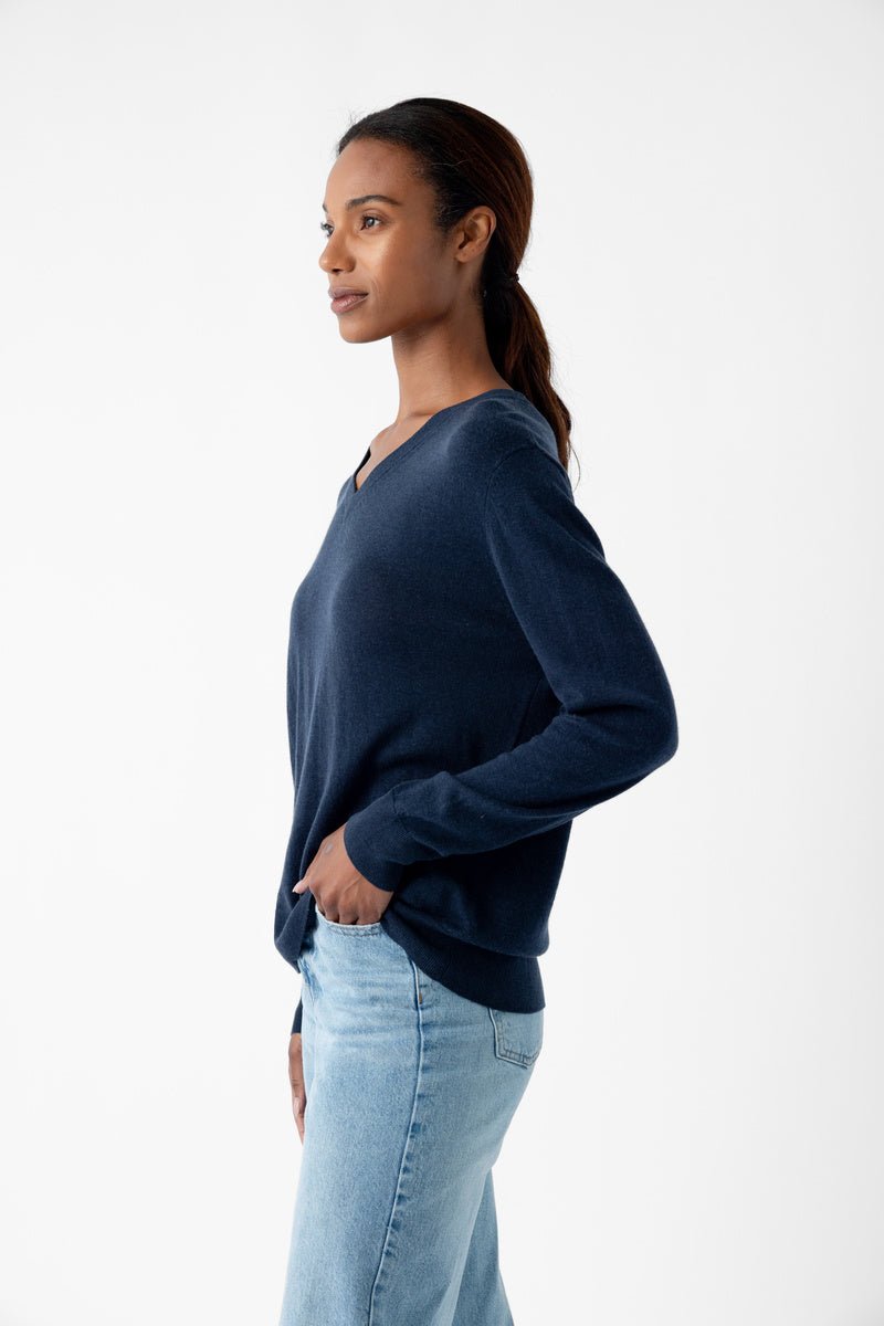 A woman stands sideways, hands in pockets, wearing Cozy Earth's Women's AirKnit V-Neck Sweater in navy and light blue jeans against a plain white backdrop. 