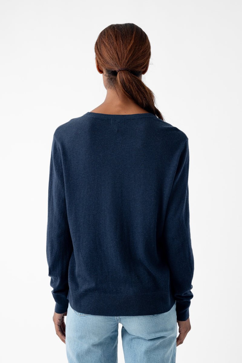 A person with long hair in a ponytail is wearing Cozy Earth's Women's AirKnit V-Neck Sweater in navy blue and light blue jeans, standing with their back to the camera against a plain white background. 