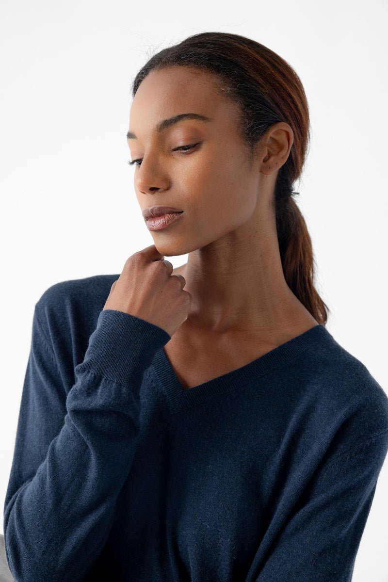 A person with long hair is looking down thoughtfully, wearing a Cozy Earth Women's AirKnit V-Neck Sweater and resting their hand gently on their chin against a plain, soft white background. 