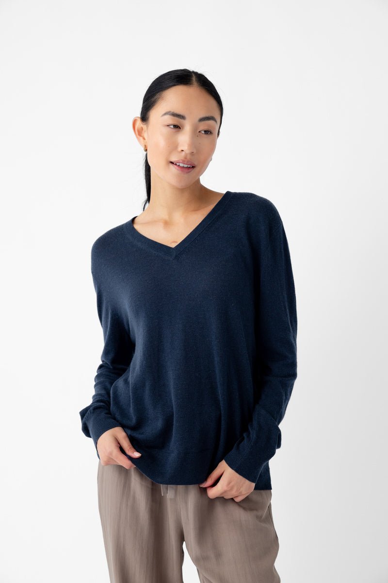 A person wearing a Cozy Earth Women's AirKnit V-Neck Sweater in dark blue and beige pants stands against a white background. They have long black hair, are looking slightly to the side with a relaxed posture, and have a subtle smile. 