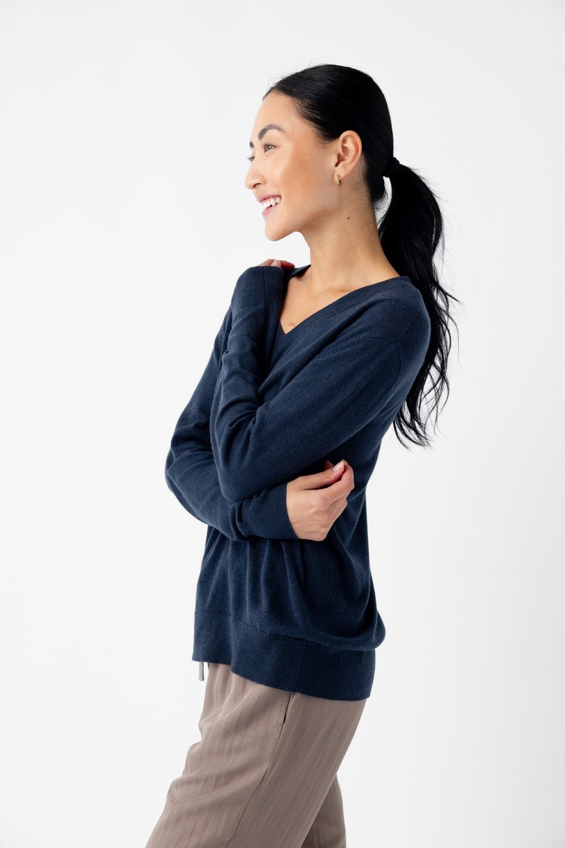 A person with long dark hair in a ponytail, smiling and standing sideways, is wearing a Cozy Earth's Women's AirKnit V-Neck Sweater in dark blue and light brown pants. They have their arms crossed and are looking off to the side against a plain white background. 