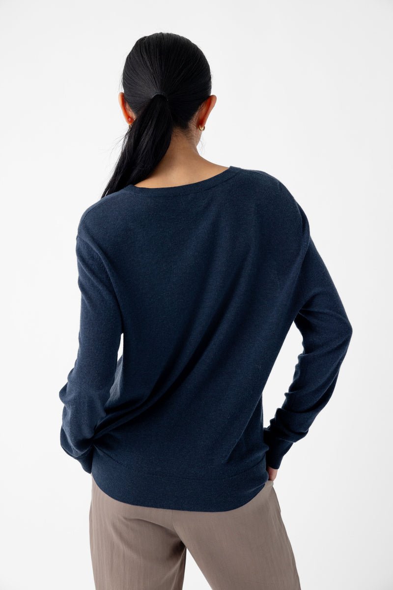 A person with long black hair in a ponytail is facing away, wearing a dark blue Women's AirKnit V-Neck Sweater from Cozy Earth and light brown pants against a plain white background. 