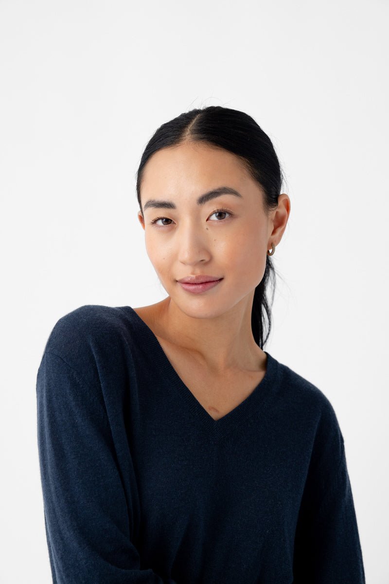 A person with long dark hair is smiling slightly at the camera against a plain white background, wearing a Cozy Earth Women's AirKnit V-Neck Sweater in dark blue. 