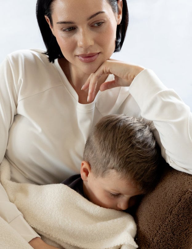 A woman in a white shirt sits on a couch with a thoughtful expression, resting her chin on her hand. A young boy sleeps peacefully beside her, wrapped in a soft white blanket.
