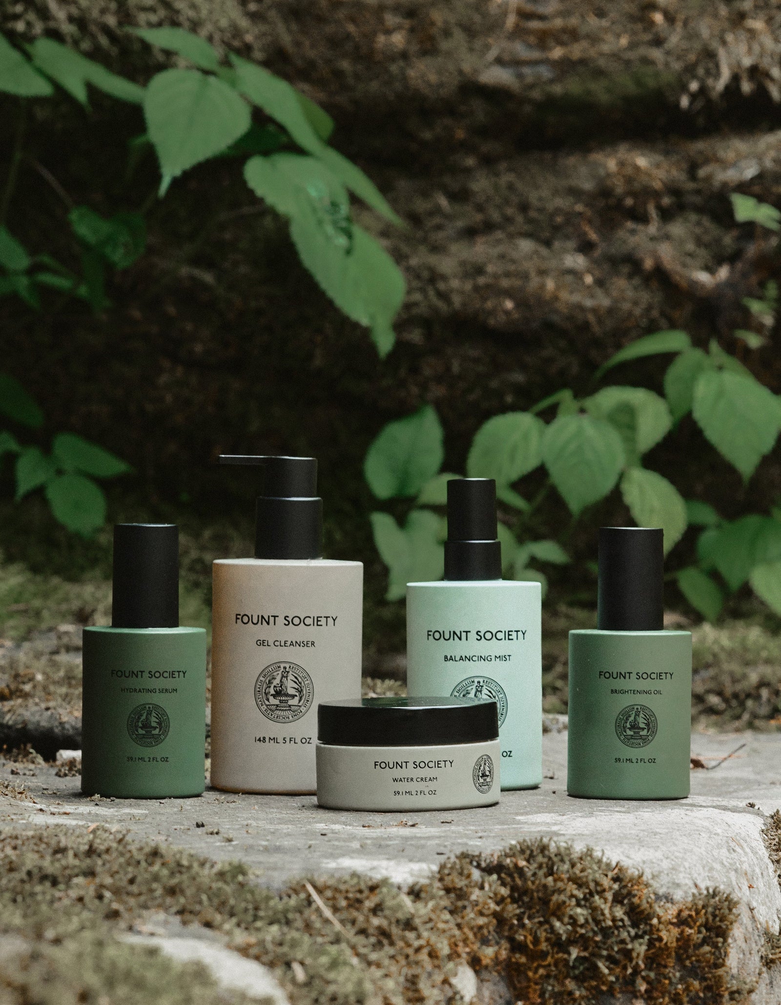 Five skincare products from Cozy Earth's "The Routine" collection are elegantly displayed outdoors on a rocky surface with lush green foliage in the background. The collection includes a gel cleanser, balancing mist, and various creams, all presented in minimalist, earth-toned packaging.