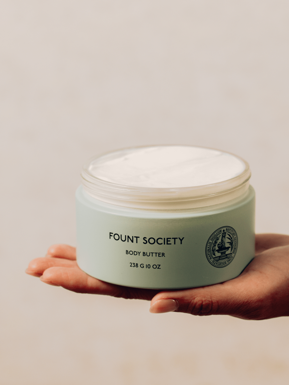 A hand holding a light green container of Cozy Earth Body Butter with a slightly opened lid, revealing the creamy product inside. The container has a 238 g (10 oz) label and a circular emblem. The background is softly blurred.