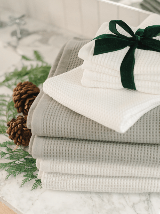 A stack of the Cozy Earth Complete Waffle Bath Bundle in beige and white rests elegantly on a marble surface, adorned with a dark green ribbon. Pinecones and green foliage beside the towels enhance the serene, natural atmosphere.