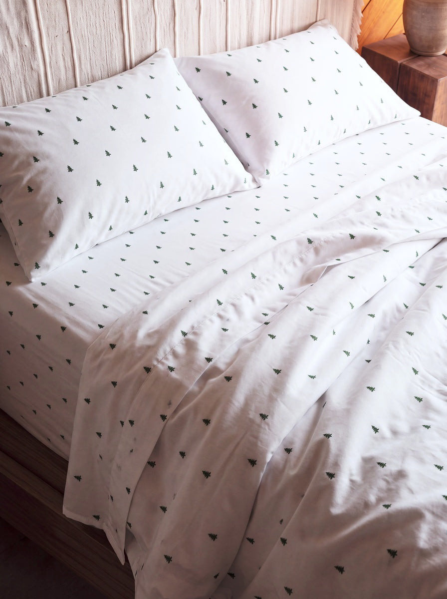 White bedding set with a small green tree pattern, including a duvet cover and two pillows, styled on a bed against a light wood-paneled wall.