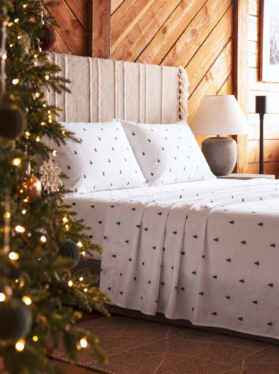 White bedding set with a small green pine tree pattern, styled on a bed in a rustic wood-paneled bedroom with a lit Christmas tree in the foreground.