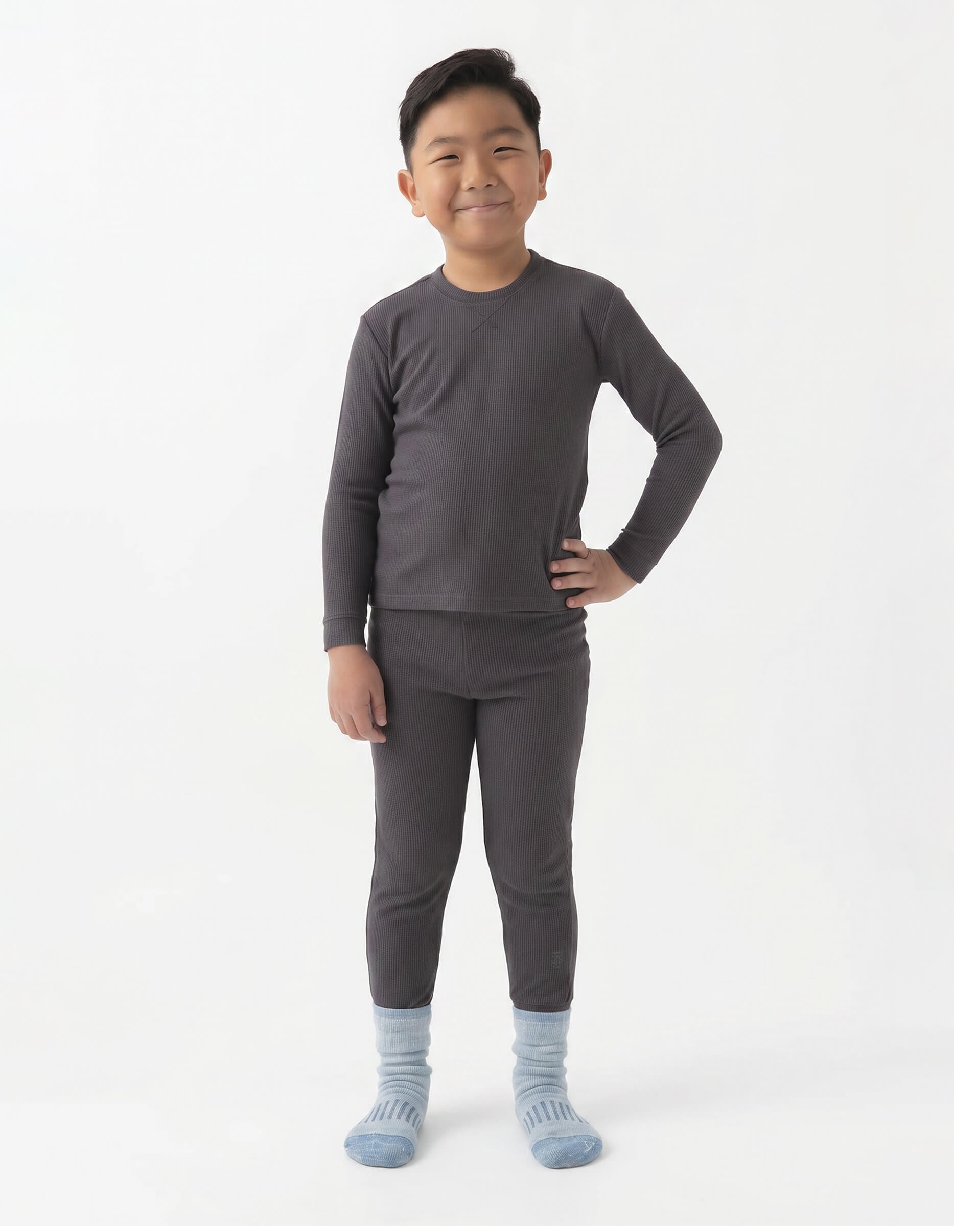 A young boy smiles with one hand on his hip, wearing the Cozy Earth Children's Bamboo Waffle Knit Base Layer Set in dark gray, with light blue socks, standing against a plain white background.