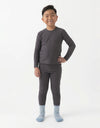 A young boy smiles with one hand on his hip, wearing the Cozy Earth Children's Bamboo Waffle Knit Base Layer Set in dark gray, with light blue socks, standing against a plain white background.
