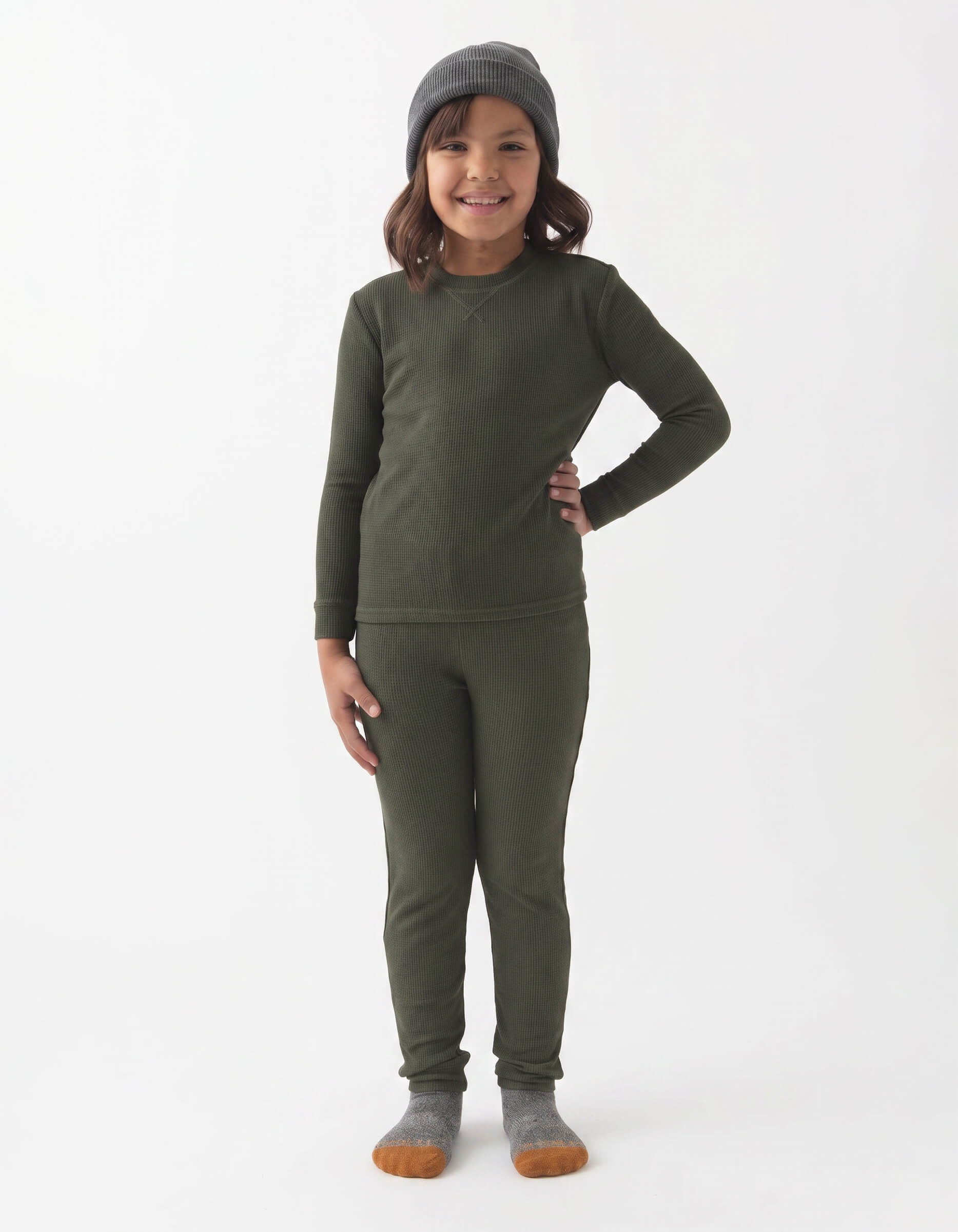 A young child smiles against a white background, wearing Cozy Earth's Children's Bamboo Waffle Knit Base Layer Set in dark green, paired with a gray beanie and gray socks with brown toes.
