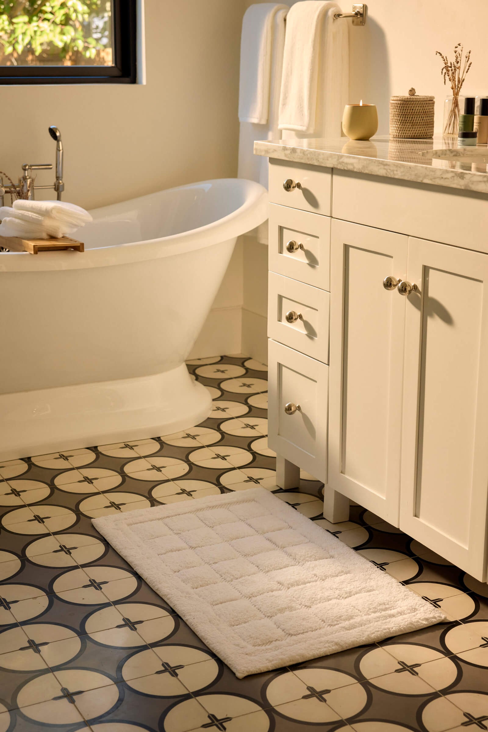 A bathroom with a patterned tile floor, a white freestanding bathtub, a white vanity with drawers, Cozy Earth's Grid Bath Mat, and neatly folded towels on the counter and towel bar. Sunlight filters in from a window.
