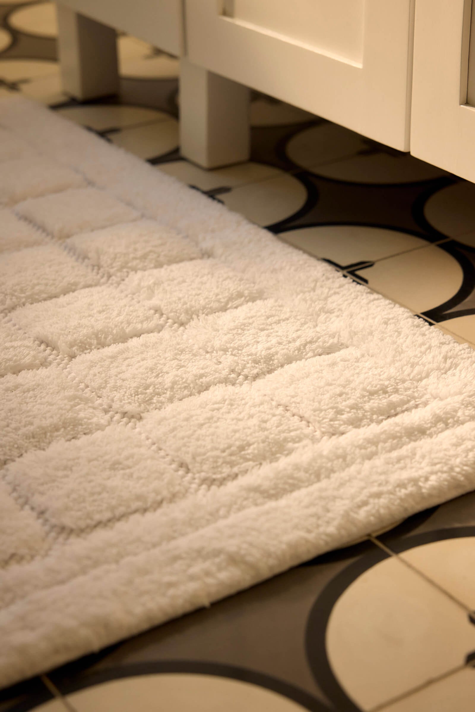 A close-up of the Cozy Earth Grid Bath Mat, featuring a white textured square pattern, placed on a circular black and white tiled floor next to a white cabinet.