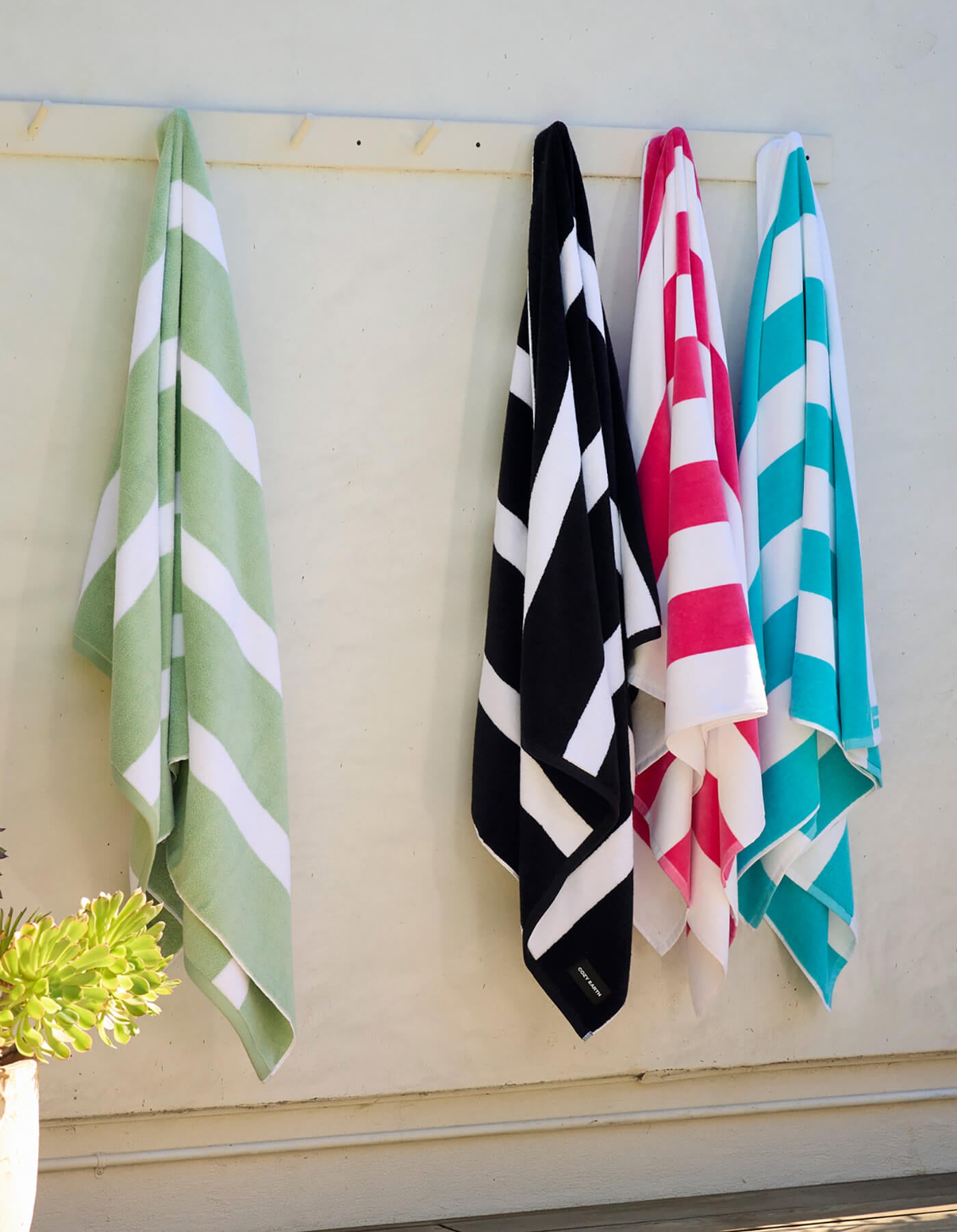 Four Cozy Earth Seaside Resort Towels in green, black, pink, and blue hang side by side on wall hooks. A potted succulent is visible in the lower left corner of the image.
