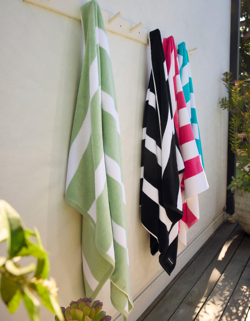 Four Classic Stripe Resort Towels by Cozy Earth in green, black, pink, and blue hang on hooks against an outdoor wall, with nearby plants and sunlight.