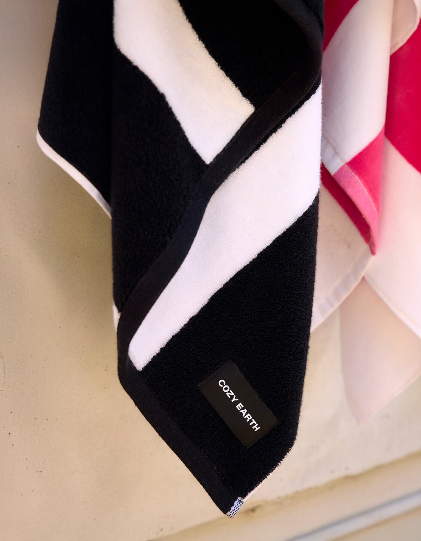 A close-up of Cozy Earth's Classic Stripe Resort Towel in black and white stripes hangs next to a pink and white towel against a light-colored wall.
