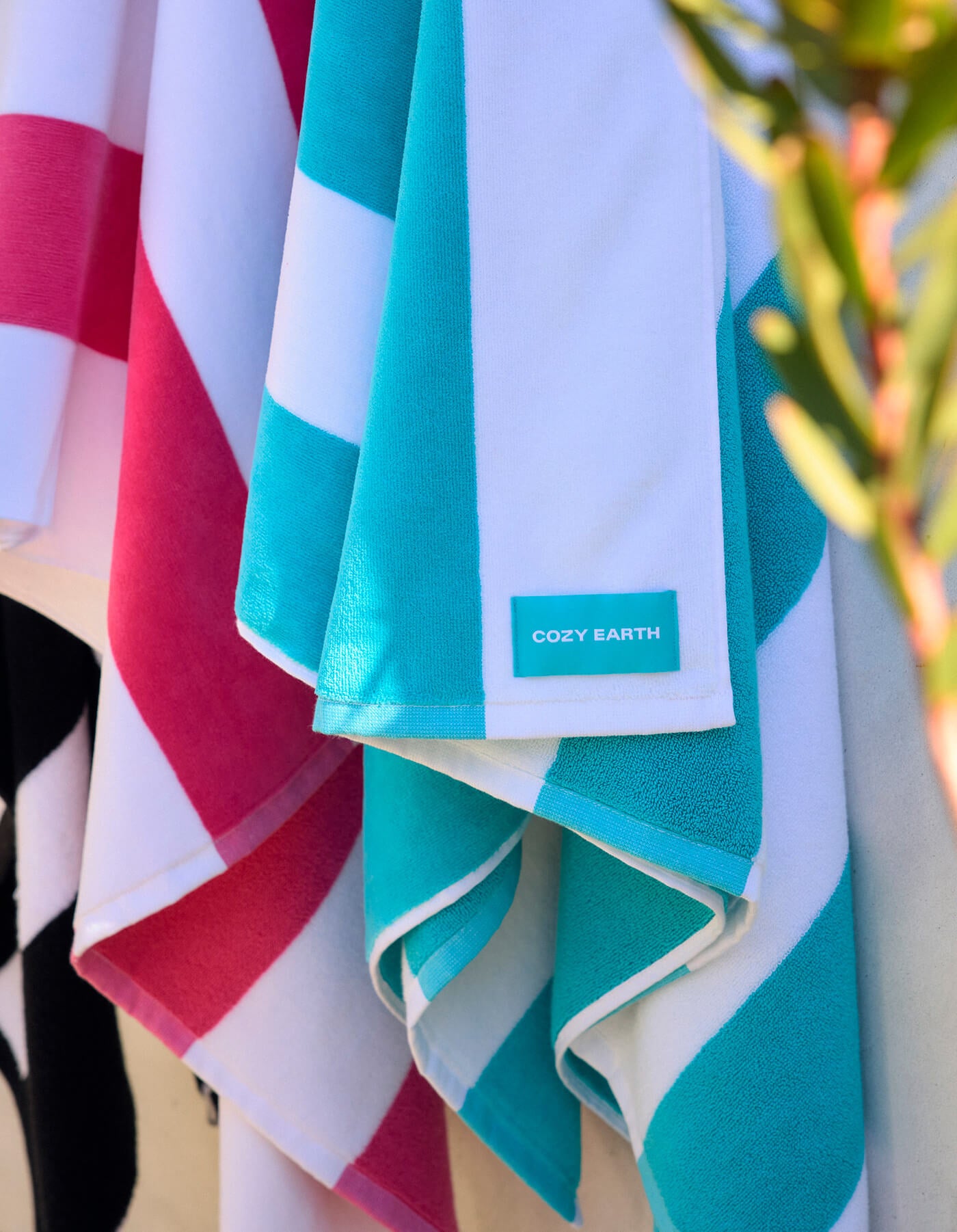 Close-up of Seaside Resort Towels by Cozy Earth hanging side by side in blue, white, and pink stripes. A label reads "Cozy Earth," and a plant branch appears on the right side.